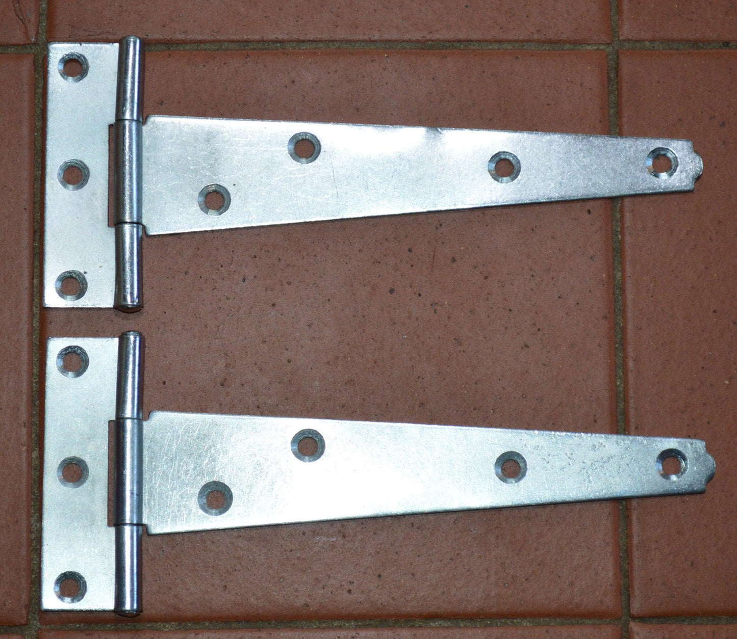 Pair of 6-inch zinc plated light duty tee hinges on brown tiled surface for sheds and gates